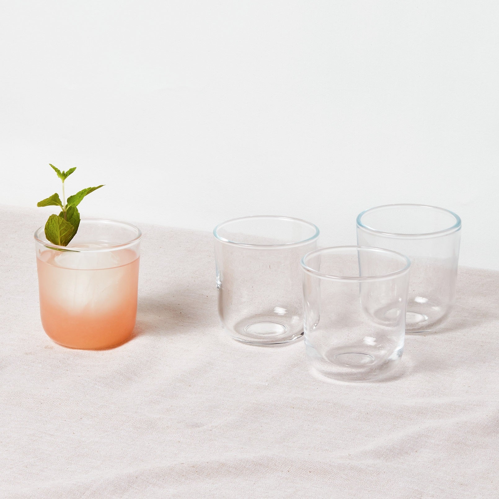 4 short glasses sitting on beige tablecloth. One glass has a pink cocktail in it and the other 3 glasses are empty.